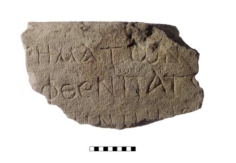 A marble fragment with a flat top and a rounded bottom, with three lines of Greek lettering. Between the first and second line, there are smaller fragments of text.