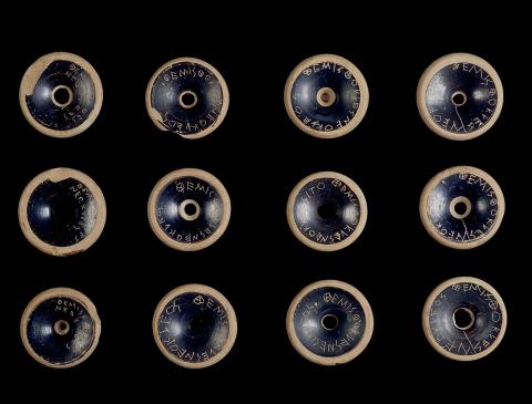 A series of 12 circular clay objects with black glaze, each inscribed with the same line of Greek lettering.