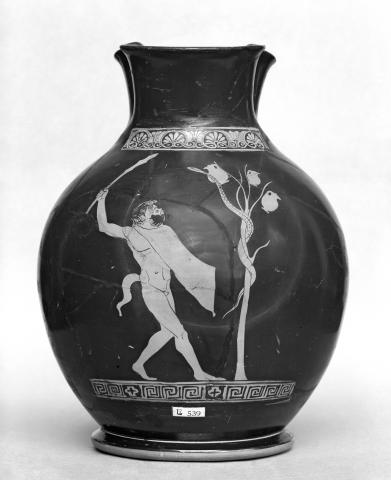 Black and white photo of the front of a jug. A satyr (left) stands in profile facing a sapling (right) around which a snake is wrapped. Three wine jugs have sprouted from the upper branches. The satyr is nude except for a wineskin cloak draped over his left arm. His right arm is raised, holding a club. He has shaggy hair and a beard. His ears are pointed and he has a tail. 