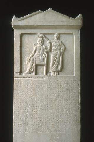 A large, long, rectabgular slab of marble, the top third of which depicts a man and woman, the rest of which contains lines of Greek lettering 