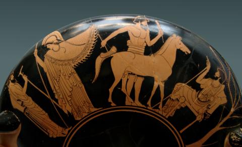 Athena (left) wearing her helmet and carrying her spear facing out turns her head toward a bearded man standing at her right shoulder. She gestures with her left hand toward a craftsman standing behind a model of a horse (center). The man and horse face right toward a third bearded man (right) seated on a stool. The craftsman is bare chested wearing a hip wrap. The other two men wear long chitons and cloaks. The craftsman holds some sort of tool in each hand.