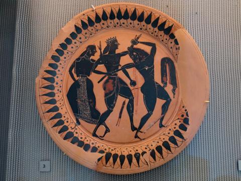 Attic Black-figure plate featuring Theseus in the center, facing right, wears a loincloth and a wreath on his head, he holds a sword in in his right hand. He is stabbing the Minotaur in the neck. The Minotaur is facing right with his head turned back toward Theseus; he holds a rock in each hand. At the left, Ariadne watches. Her body faces left and her head turns right, toward Theseus. Her left arm is raised, gesturing. The plate's rim is decorated with a design of lotus blossoms.