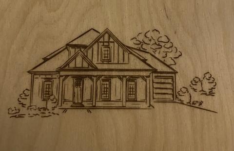 Line drawn woodcut of a house surrounded by bushes and trees. A porch extends across the front of the house supported by wooden beams. A mat sits on the porch in front of the door. 