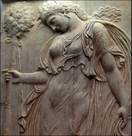 A maenad dances holding a thyrsus in her right hand. She wears a very light, almost transparent, dress, earrings and a what appears to be a diadem. Her hair is down, but gathered in a loose, low ponytail.