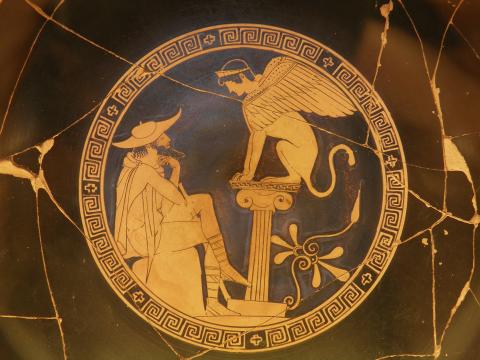 A bearded Oedipus, dressed in a traveler's hat and cloak sits on a rock, left. His legs and arms are crossed, his chin resting on the back of his left hand. A sword or walking stick rests against his legs. He stares at the Sphinx, right, who sits on a short pillar. Her(?) wings are swept back against her cat body and slightly raised. She wears a crown or diadem on her head. A circular meander encloses the scene.