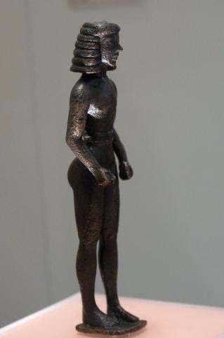 Right profile view of a nude male youth. His hair is molded in 6 horizontal lines, reminiscent of a wig. He wears a belt at his waist. His arms are held close to his sides, slightly bent at the elbows. His hands are in fists. He stands with his left foot slightly forward.