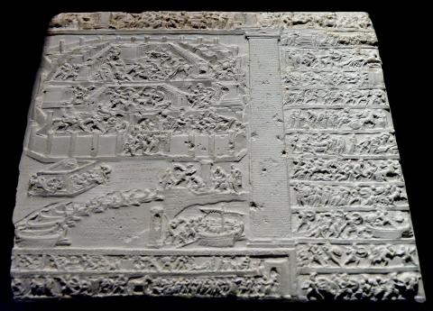 Rectangular fragment of a relief carving. Two-thirds of the fragment is a single large image of the fall of Troy. In the foreground, the beached ships of the Greeks. In the background Troy, with the main gate open. Fighting rages throughout the streets, agora, and temple precinct. Legends in Greek, carved in the negative space of the beach and harbor, describe the scene. Horizontal bands of battle scenes border the scene above, below, and to the right of the image. 