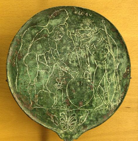 Back of a bronze mirror, the metal has formed a green patina. Etched on it are the figures of two men. Left, a nude young man, his left foot resting on a rock, leans forward to grasp the handle of an amphora with his right hand. Across from him an older bearded man wearing a chlamys drinks from a double-handled cup in his left hand. In his right, he holds a staff or spear. A small fountain sits between them.
