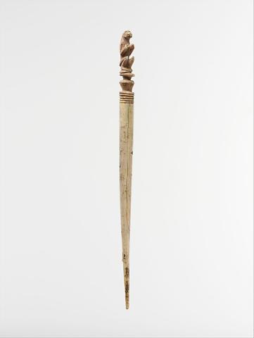 Ivory hair pin. One end is tapered to a point to be inserted in the hair. The opposite end features a carving of a monkey sitting on his back haunches with his front paws raised at chest height. The monkey carving sits atop a turned section of ivory shaped like a small pedestal, below which the ivory is incised in five concentric bands.