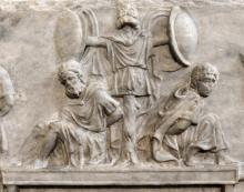 Marble relief panel from the Arch of Titus. In the center of the image are two men sitting on a large platform or litter with their hands bound behind their backs. The men are barefoot wearing long tunics and cloaks consistent with Roman depictions of Gauls, Germans and other men from the northern frontier. The men are seated at the foot of a wooden frame constructed from a tree trunk with crossbars. Four round shields and clothes similar to the captives are hung on it. 