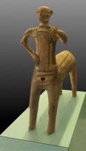 Terracotta centaur, the horse body is wheel-made with a small hole in the chest of the horse for ventilation during firing. The human portion is handmade. Reddish-brown paint decorates the horse back and human chest and arms with geometric patterns. The left arm is missing just below the bicep. The right arm is bent with the six-fingered hand resting on his hip. There is a vertical gash on the front left leg at approximately the knee. The head was broken off and reattached.
