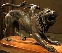 Three-quarter view of Chimera facing right. It's in a half-crouch, front legs outstretched. The lion's mouth is open as if roaring. A goat's head protrudes from the animal's left flank. It is turned away from the viewer and leaning to the side. Drops of blood flow from its neck. The lion's tail is a snake, curved up and over the lion's back. It grasps one of the goat's horns in its mouth. The lion's mane and a line of fur down its back stand up from its body and curved barbs.
