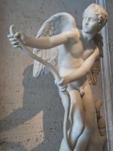 Marble statue of Cupid stringing his bow. Cupid faces front with the bow held to his right side and head turned to face it. He holds the grip with his left hand and grasps the top limb with his right.