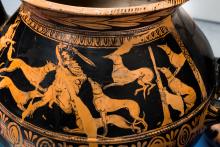 Actaeon, in mid-transformation, is surrounded by his 6 dogs, three of which are already attacking. He has sprouted the ears and antlers of a stag; his word is raised behind his head in his right hand. He is nude except for a cloak secured by a round broach at the neck. He has wrapped his it around his left arm, perhaps in defense. He is bleeding from bites to each leg and one on his torso. Artemis stands at the far left, almost out of frame