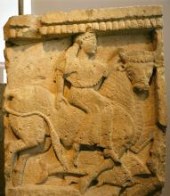 Europa, in right profile, sits sideways on the back of a bull. She wears a chiton and wrap; her hair hangs down her back, gathered at the neck. The bull's head is turned to look at the viewer. Europe grasps its right horn with her left hand. Below the bulls hooves are two dolphins.