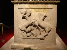 A nude youth lies on the ground between to rearing centaurs. He is visible from torso up, trying to protect himself with a shield strapped to his left arm. The right-hand centaur is broken off at the shoulders. The left-hand centaur holds a pithos over his head with both hands preparing to swing it down on the head of the youth.