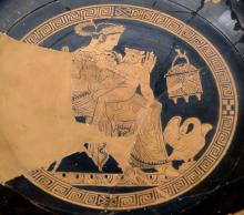 Interior of an Attic red-figure kylix showing Pasiphae seated on a chair, facing right, holding the young Minotaur on her lap. The Minotaur is painted with a miniaturized version of a fully grown man's body and a calf's head. Pasiphae appears to be dressed in a peplos and sandals. She wears pendant earings and a bracelet on her right wrist. A swan is painted at her feet, a small chest with a domed lid hangs from a hook on the wall in the background. The scene is surrounded by a circular meander.