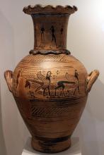 Terracotta water jug with two registers of decoration. Around the neck a chain of young men and women holding hands, painted with reddish-brown pigment in silhouette. Both have shoulder-length hair, the women wearing long skirts, the men apparently nude. On the belly, left to right, a man in silhouette followed by two sphinxes, also in silhouette, with cat bodies, wings with feathers depicted by circles. Above and below the jug is decorated with bands of digaonal wavy lines and lattice-like designs.