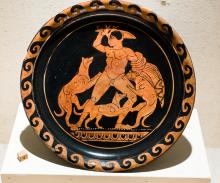 Red-figure plate showing a nude Acteon being attacked by three dogs. HIs sword is raised above his head in his right hand. His cloak is wrapped around his left forearm. He appears to grab one of the dogs around the neck with his left hand.