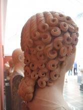 Rear view of a marble bust of a woman. The right side of the head is shown. A series of of curls, beginning about 2/3 of the way across the crown of the head, cascades diagonally from the temple along the skull toward the nape of the neck. The bottom few inches of her hair, beginning approximately where the base of the neck joins the shoulders, transitions from curls to a braid.